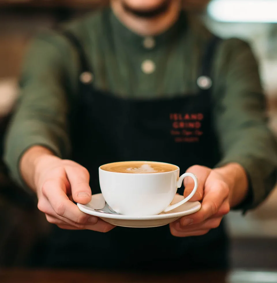 An Island Grind barista in a green apron serving a cappuccino